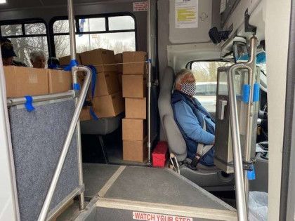 Passenger in blue jacket seated on a bus beside stacked boxes and a wheelchair area