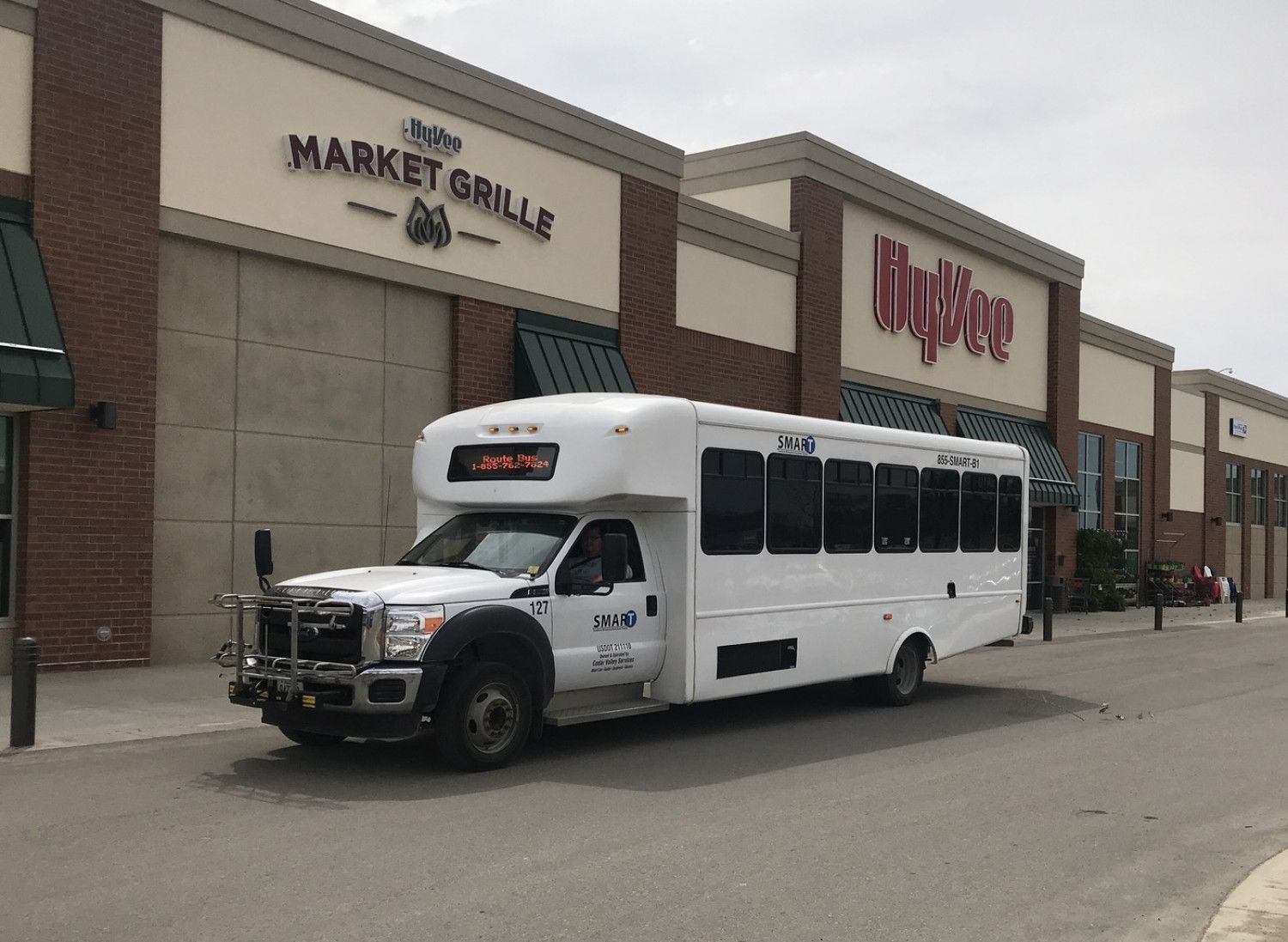 White shuttle bus parked outside Market Grill and Walgreens in a shopping plaza.