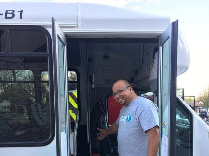 Man standing at the open door of a bus, smiling and gesturing inside.