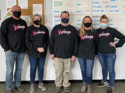 Five masked people in matching black shirts standing indoors in front of a whiteboard