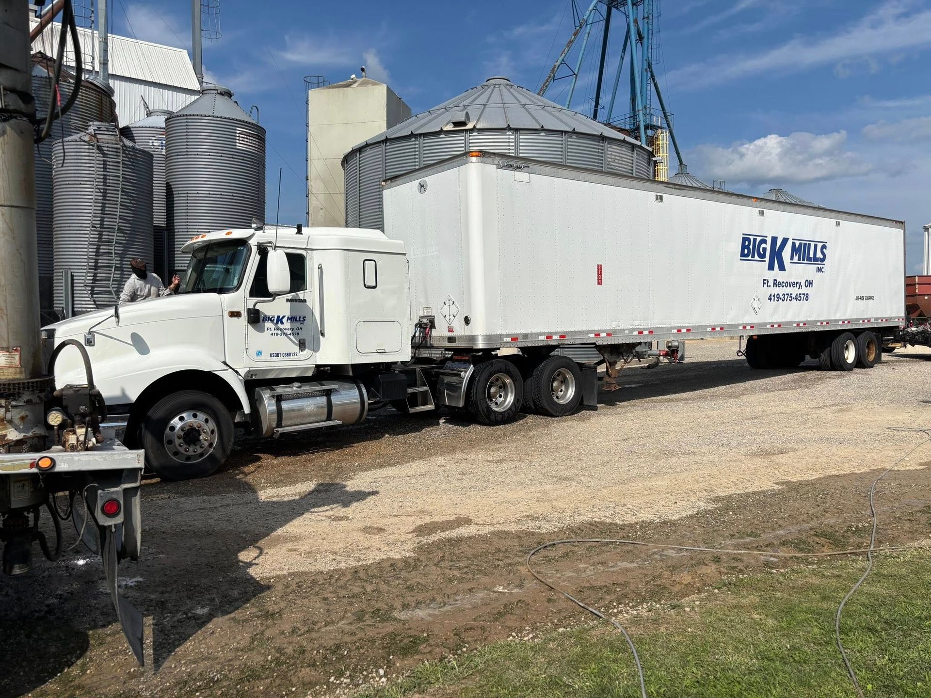 White semi-truck parked near grain silos, possibly loading, on gravel surface, blue sky in background.