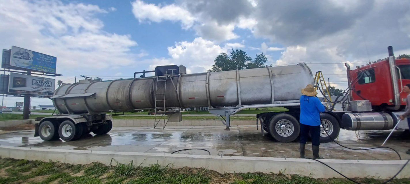 A tanker truck being washed by two people. The truck is silver and red. Outdoors with cloudy sky.
