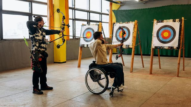 Zwei Personen beim Bogenschießen in einer Halle: eine stehend, die andere im Rollstuhl, beide zielen auf Zielscheiben.