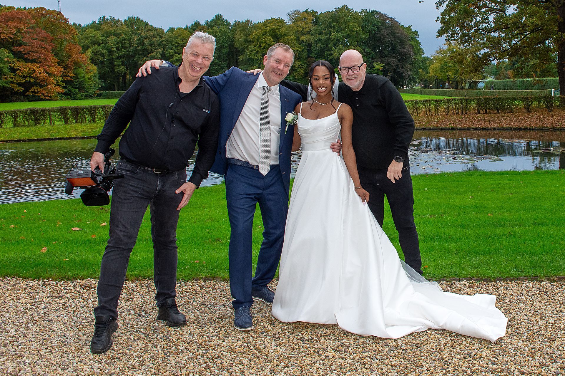 Een bruid in een witte jurk poseert met drie mannen buiten bij een vijver.