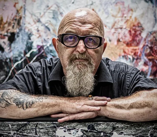 Man met baard en bril, zijn armen rustend op het oppervlak voor een kleurrijk kunstwerk.