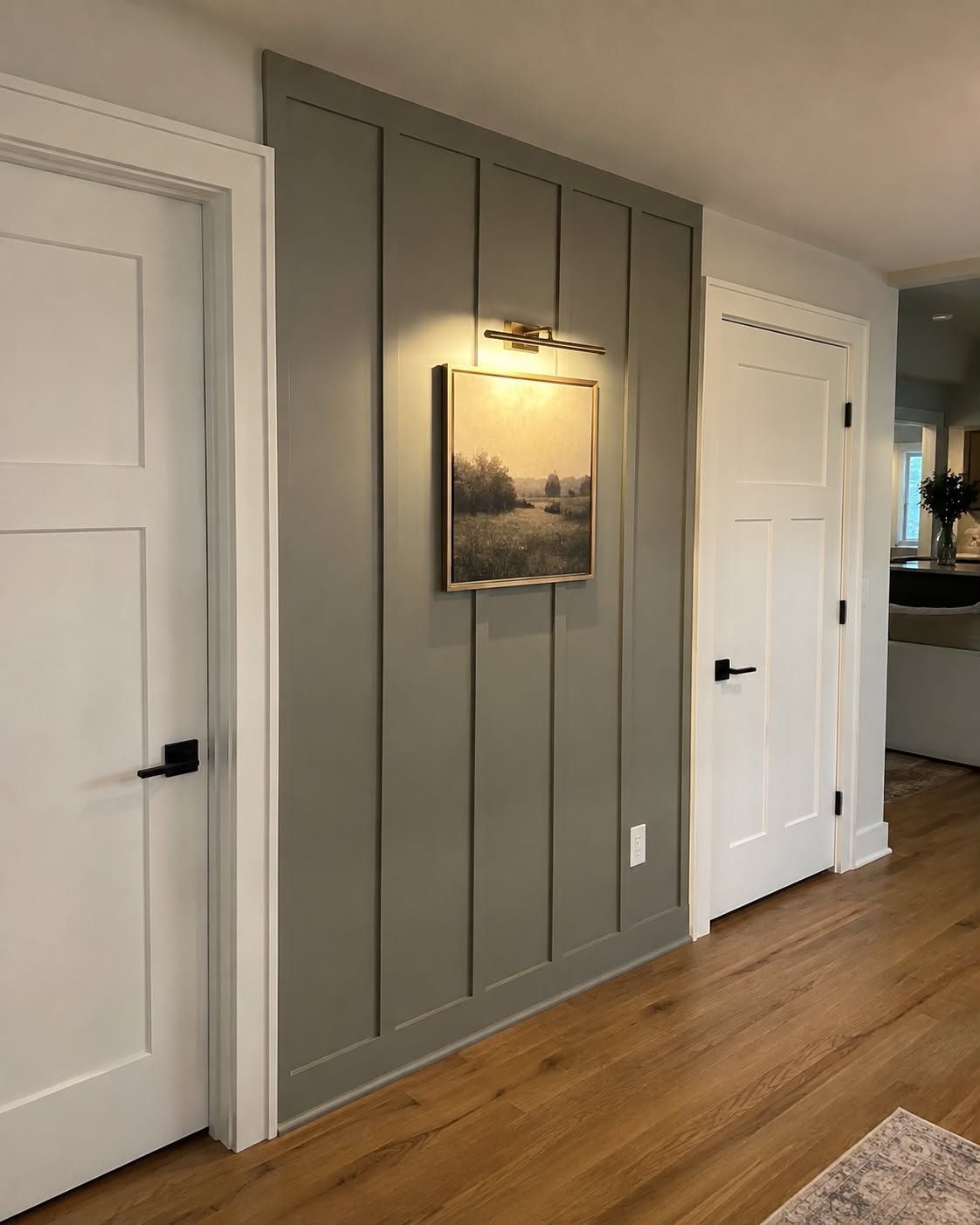 Hallway with gray paneled wall, framed art under a light, white doors, and wood flooring.