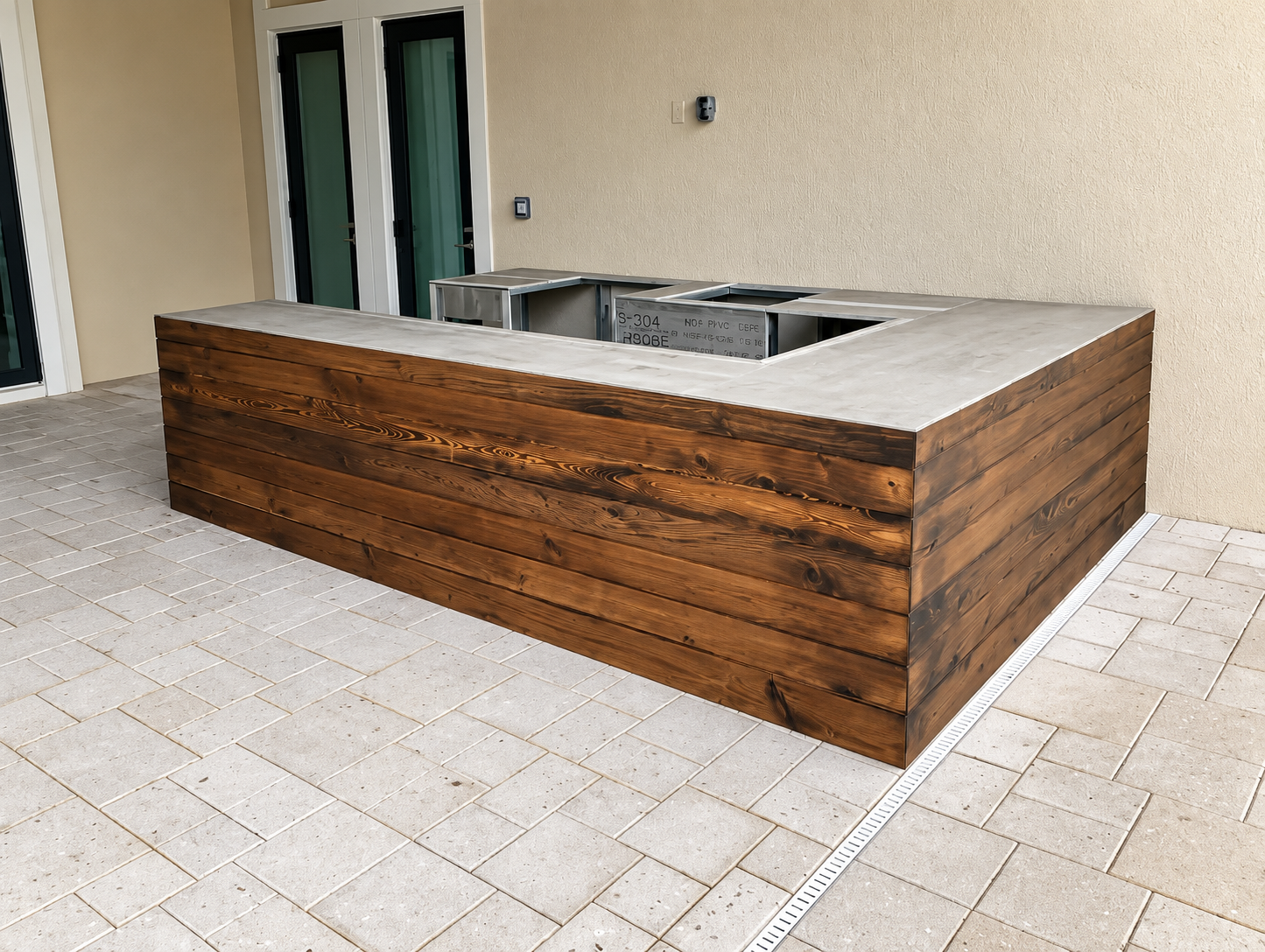 Wood-paneled outdoor kitchen island with stainless steel sink on a tiled patio near a house door