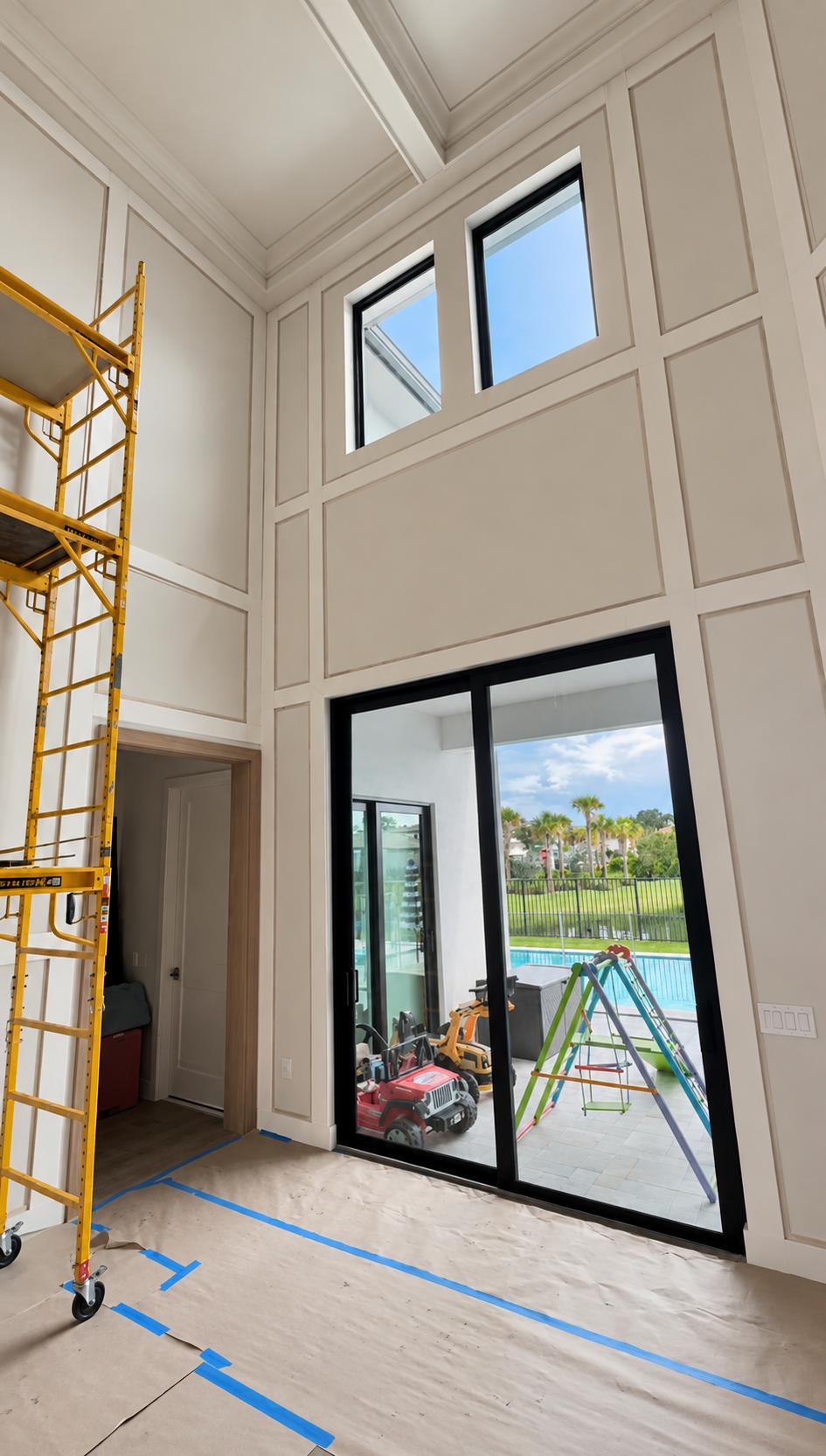 Tall unfinished interior with scaffolding, large glass doors, and blue floor tape leading to a bright patio view