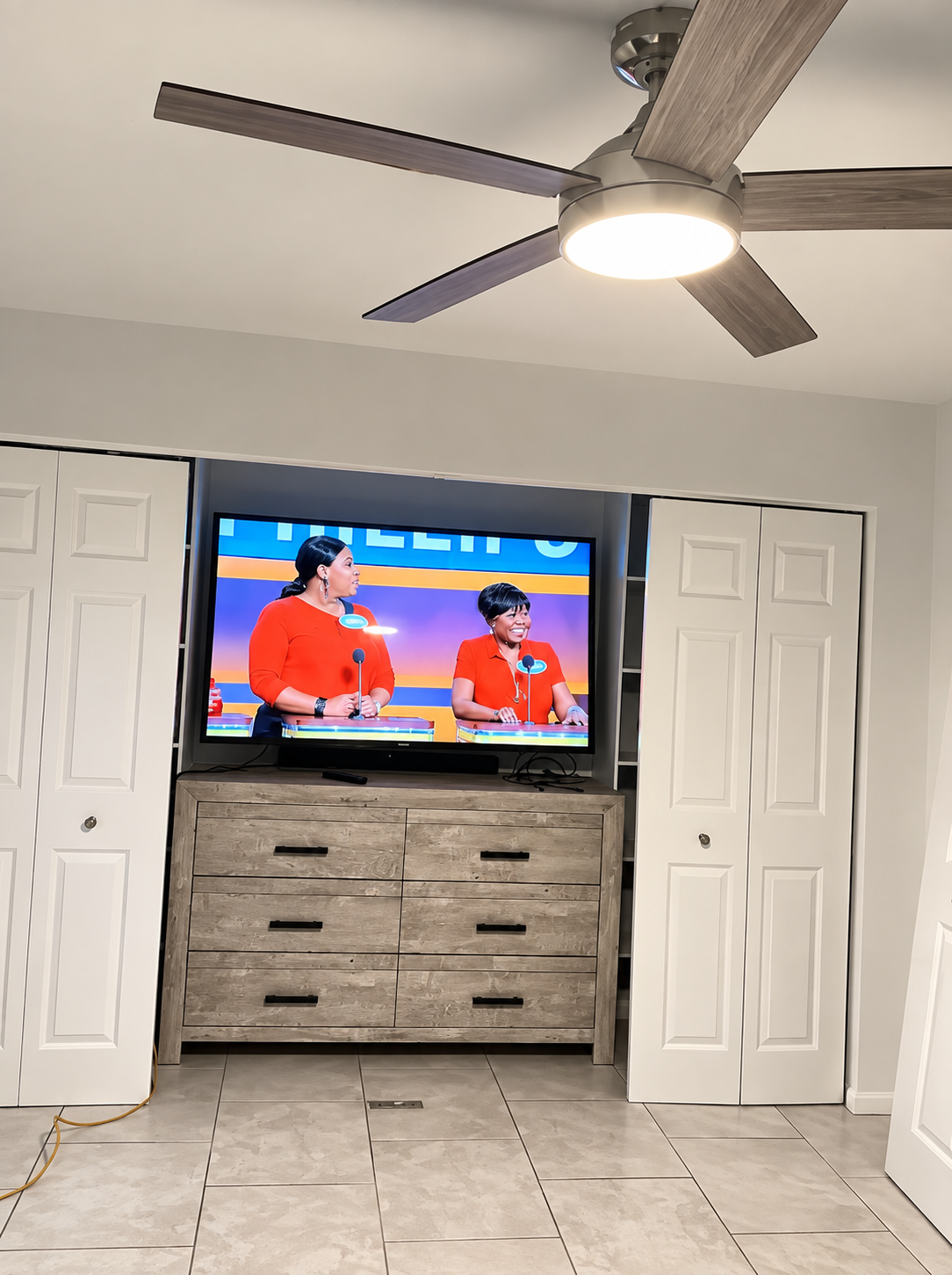 Bedroom with ceiling fan and TV above a dresser showing two people in red shirts on a broadcast.