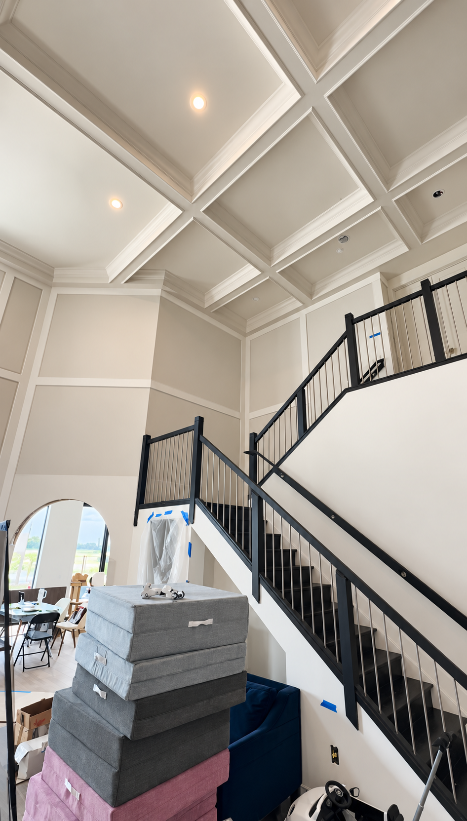Staircase in a high-ceilinged foyer with white coffered ceilings, black railings, and stacked floor tiles.