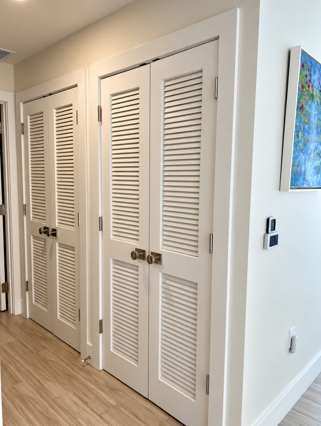 White louvered closet doors in a bright hallway with wood floors and wall art.
