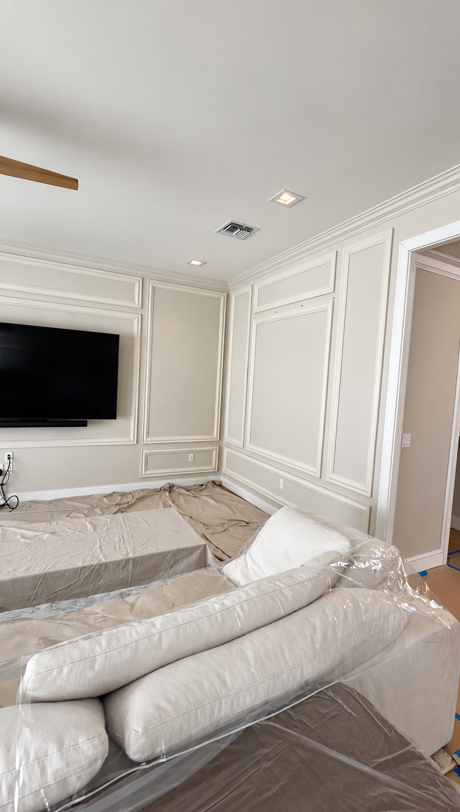 Living room with white paneled walls, wall-mounted TV, and furniture covered in protective plastic