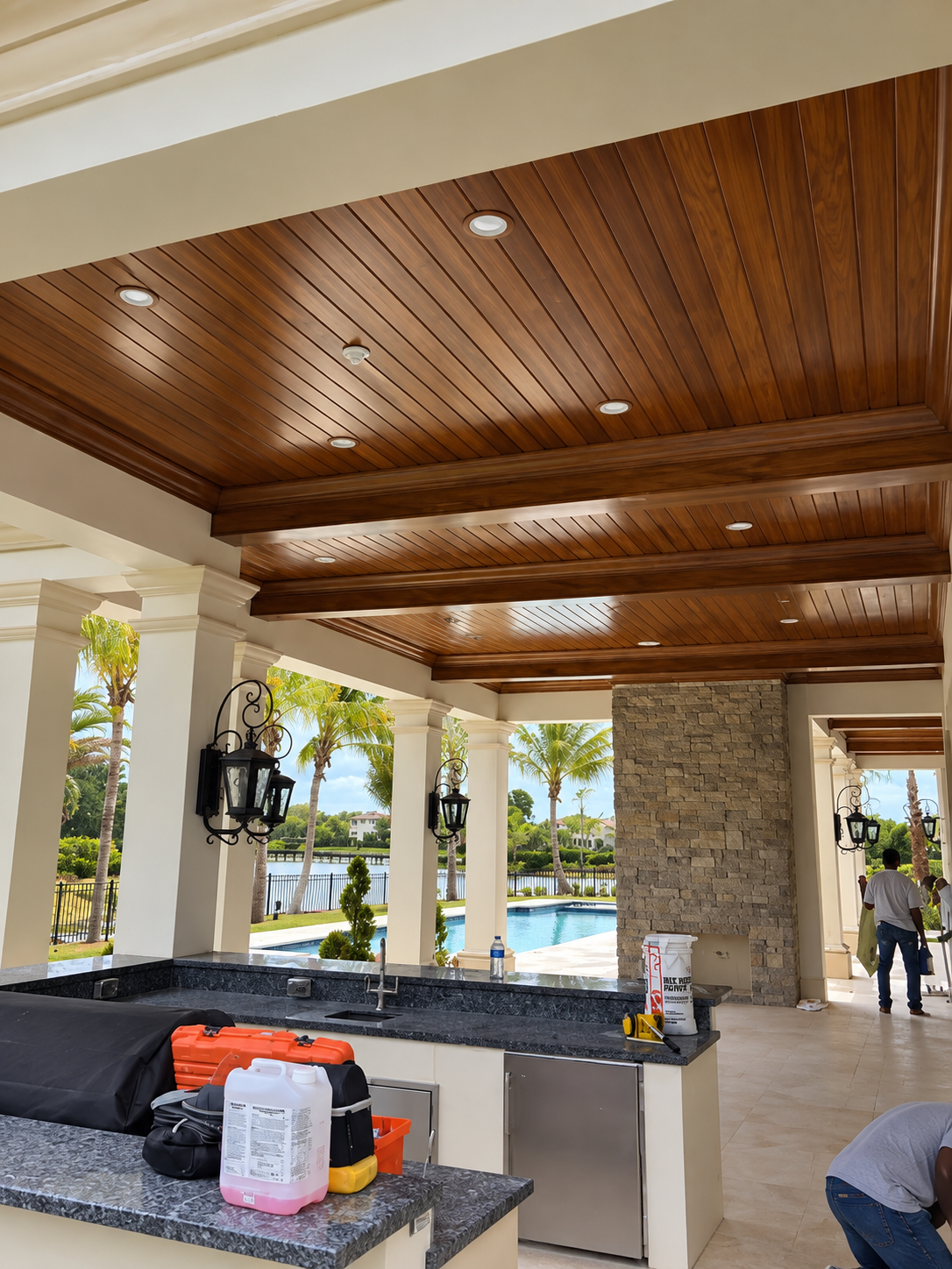 Covered outdoor bar with stone counter, wooden ceiling, and poolside view through open columns.