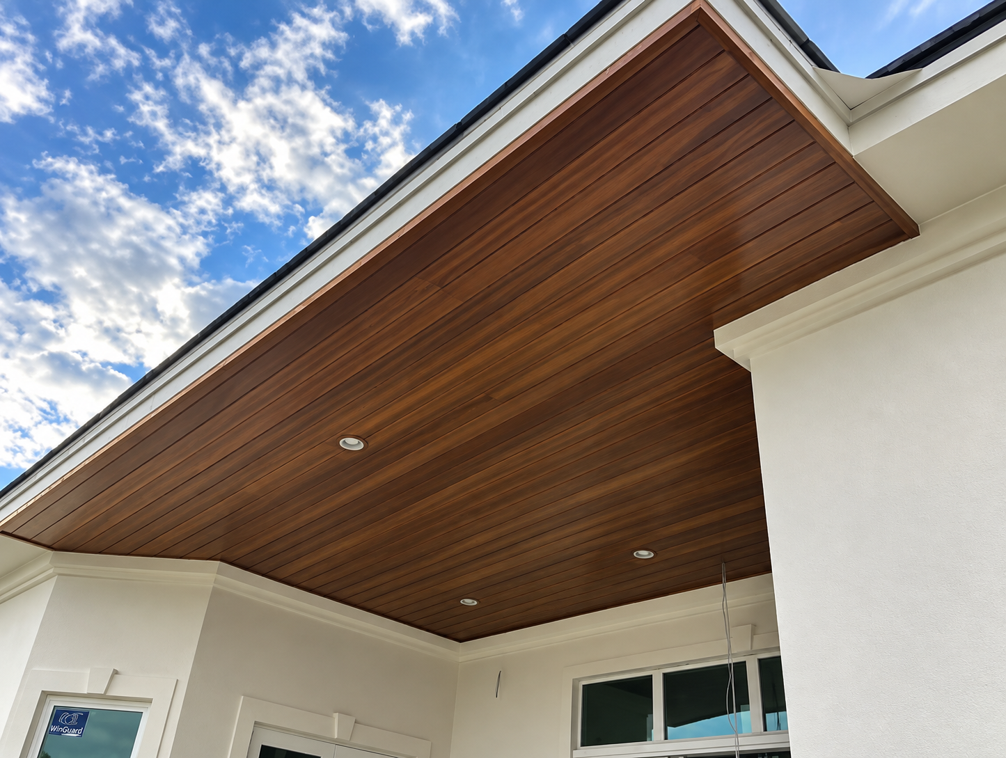 Wood-paneled ceiling over a white house porch with recessed lights and blue sky visible above