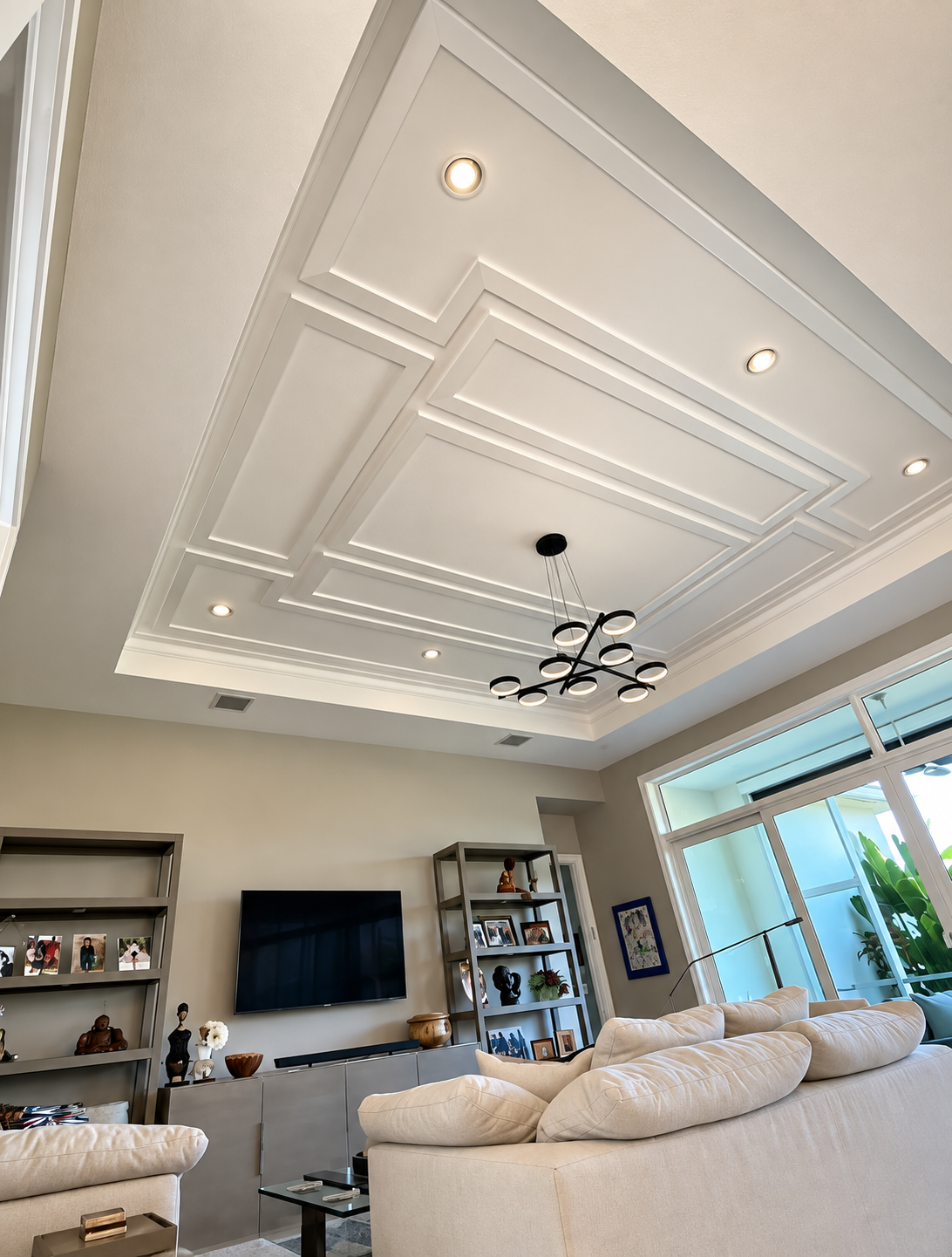 Bright living room with coffered ceiling, recessed lights, TV, shelves, and sectional sofa by large windows