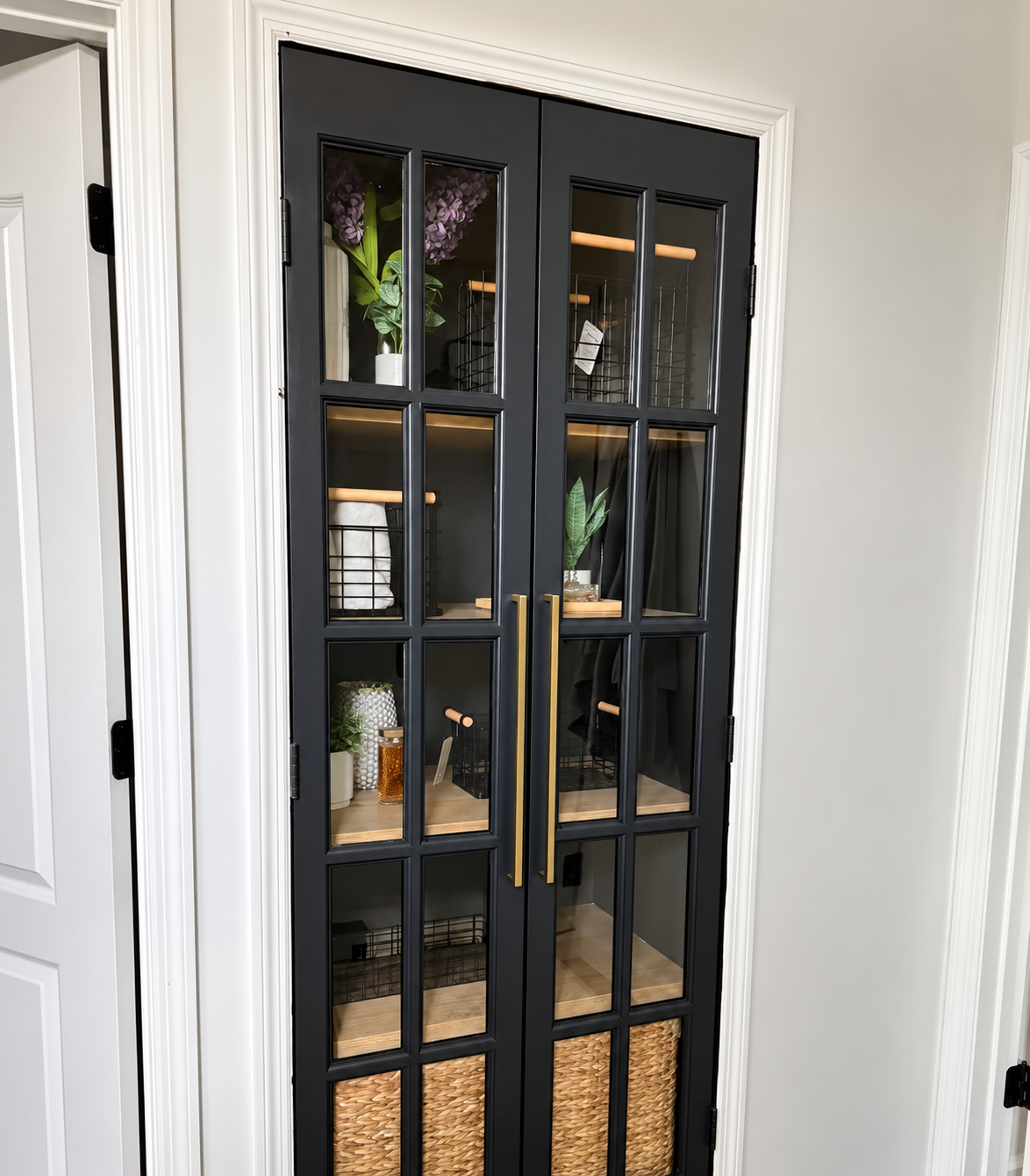 Black-framed glass cabinet with shelves holding books, decor, and woven baskets in a light room