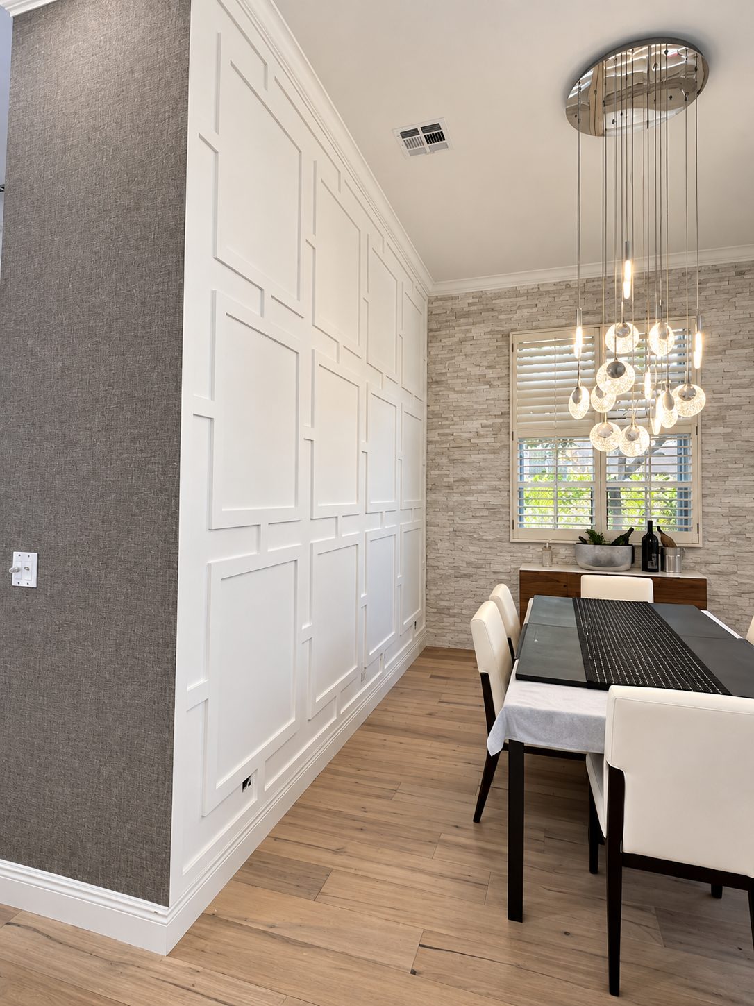 Bright dining room with white chairs, chandelier, large cabinets, and gray walls beside a window.