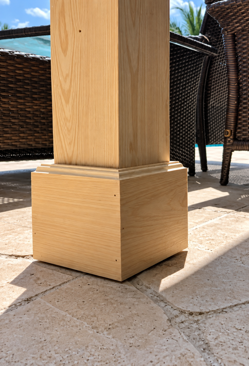 Wooden patio post on tiled floor beside wicker chair, with poolside sunlight visible behind.