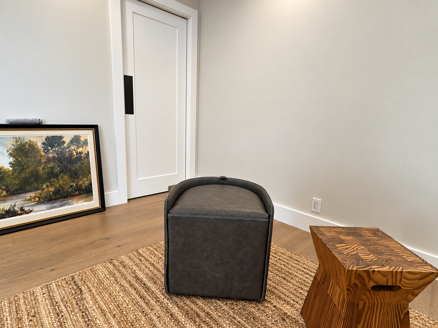 Small empty room with a framed landscape leaning against the wall, a black ottoman, and a woven basket stool.