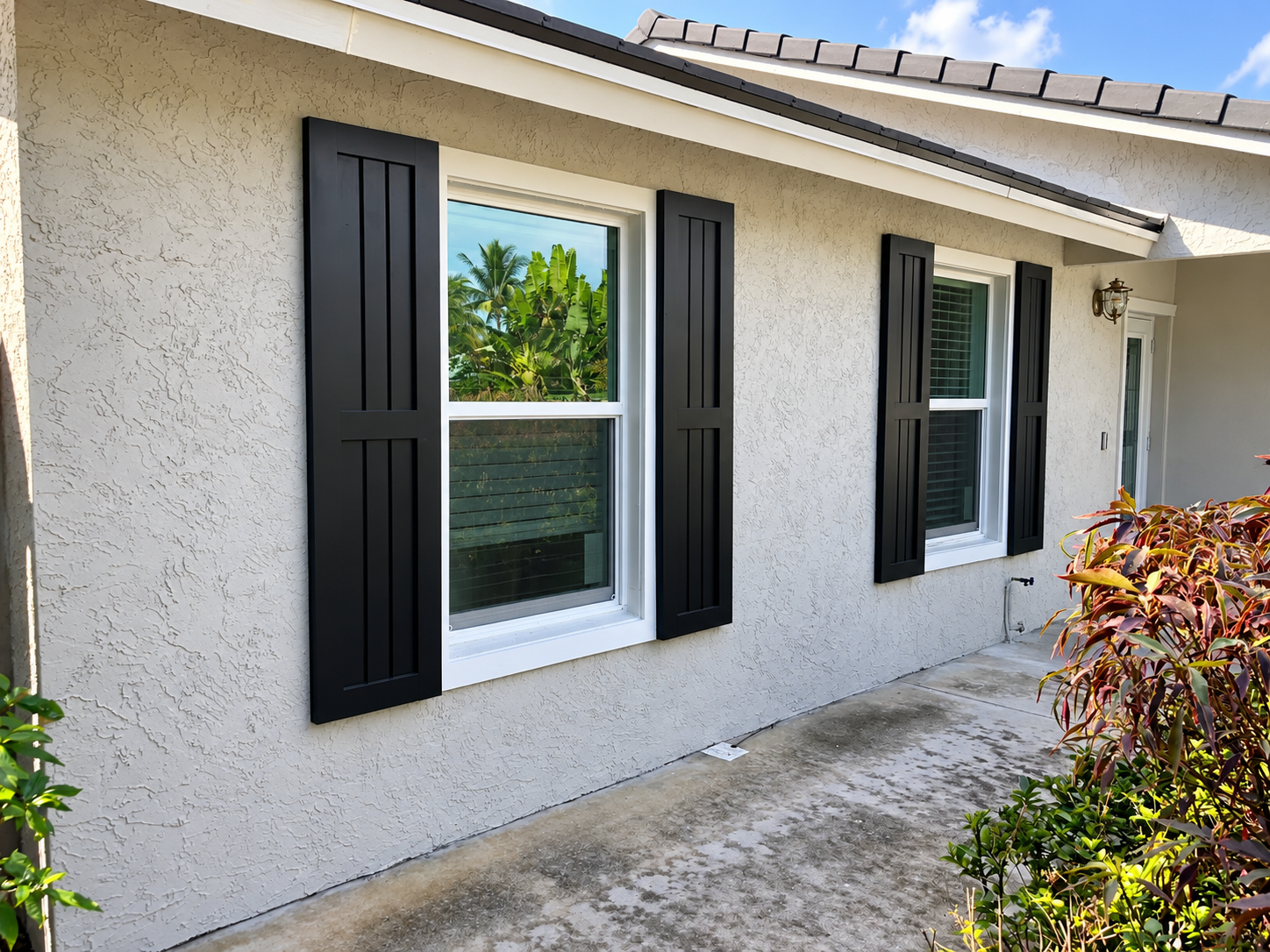 Exterior of a beige house with black shutters, white-framed windows, and a concrete walkway beside bushes