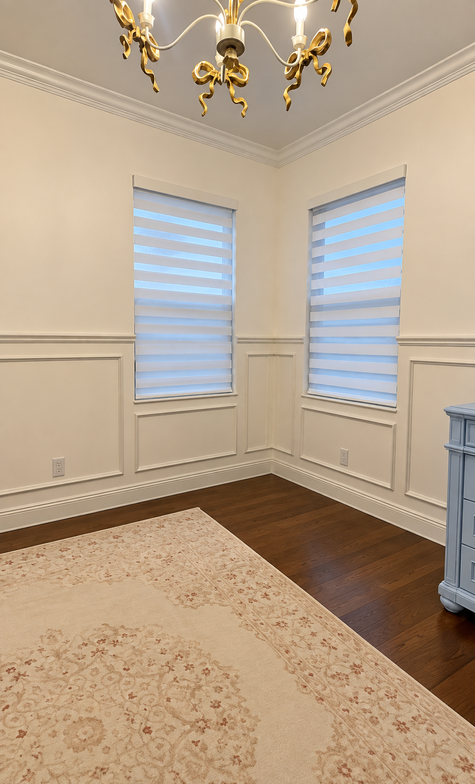 Elegant empty corner room with cream walls, bay windows, chandelier, and hardwood floor.