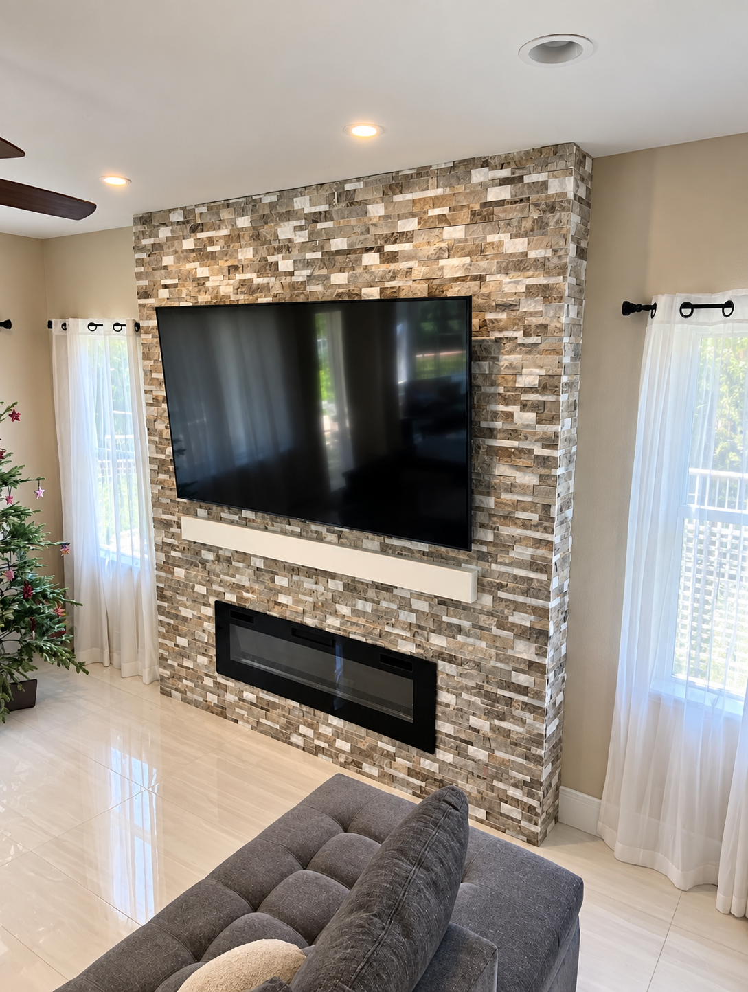 Living room with stone TV wall, wall-mounted TV, fireplace, and large windows with sheer curtains