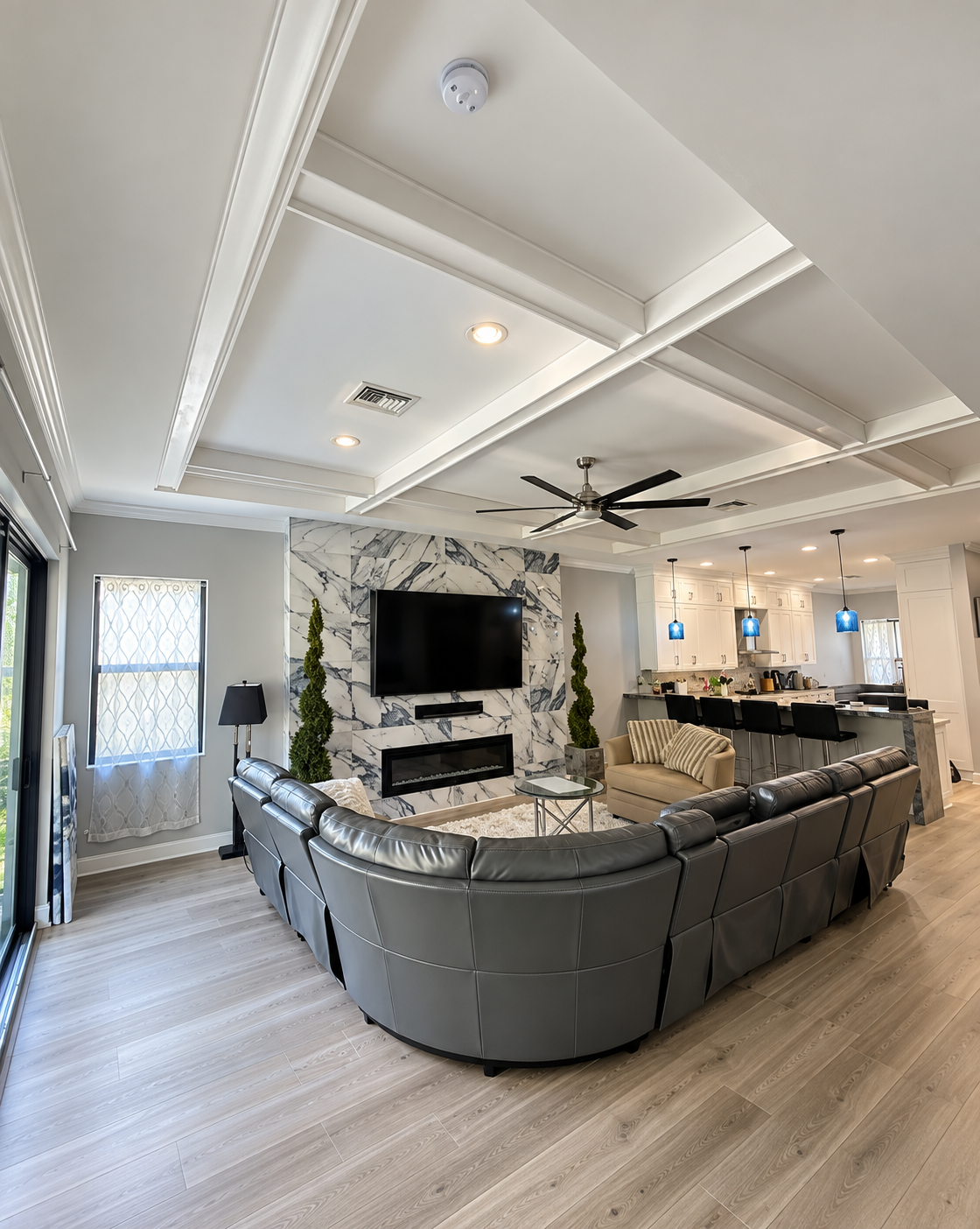 Bright modern living room with sectional sofa, wall-mounted TV, fireplace, ceiling fan, and coffered ceiling