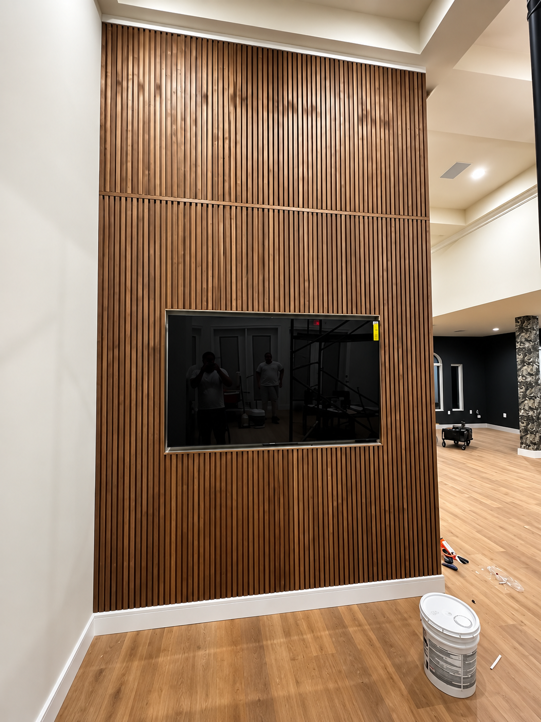 Wall-mounted TV on a wood slat accent wall in a modern room, with a paint bucket on the floor.