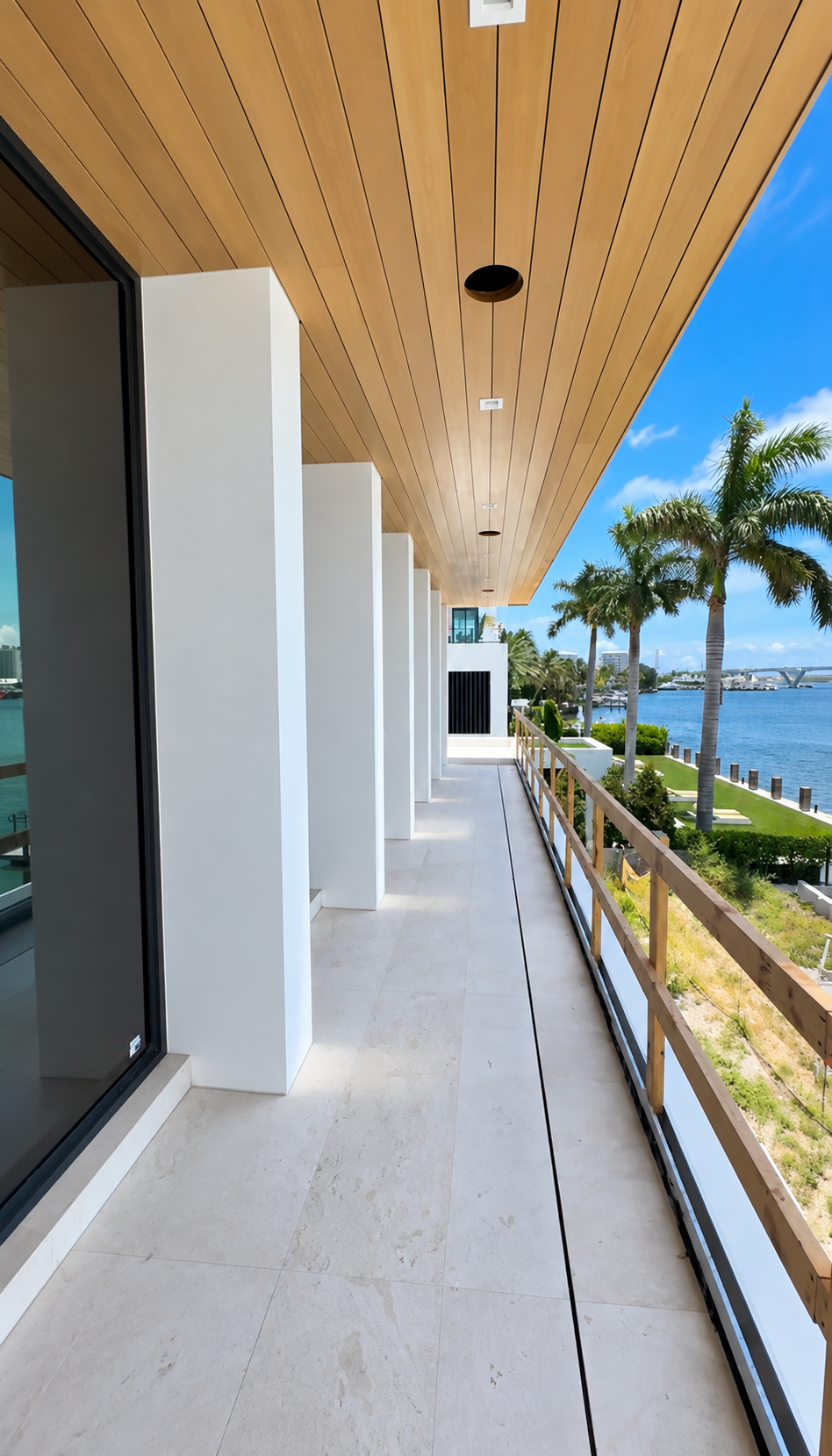 Modern covered balcony walkway with white columns, wood ceiling, and ocean view with palm trees