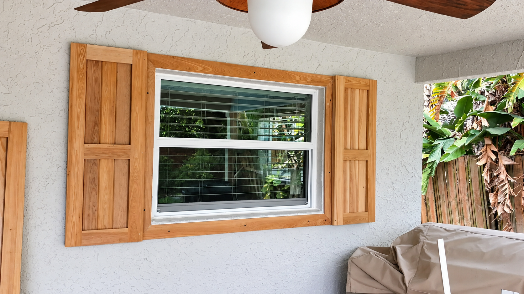 Wooden-shuttered window in a white patio wall, with a ceiling fan and leafy greenery outside.