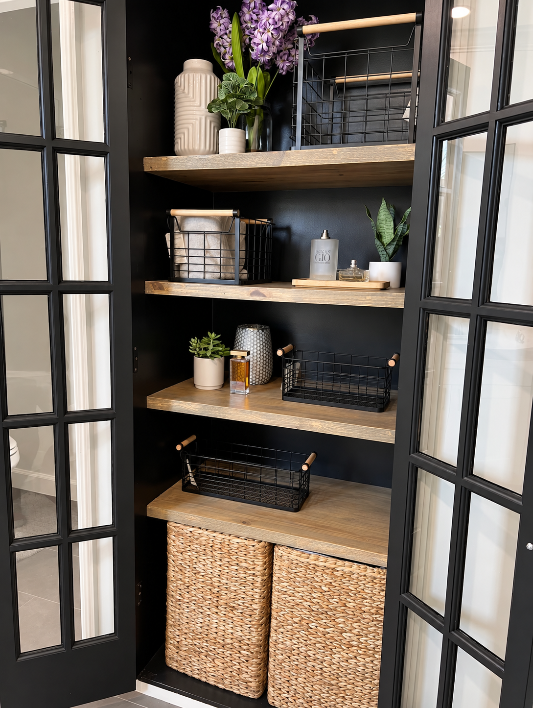 Open black-framed cabinet with wooden shelves, baskets, mugs, and potted plants on display
