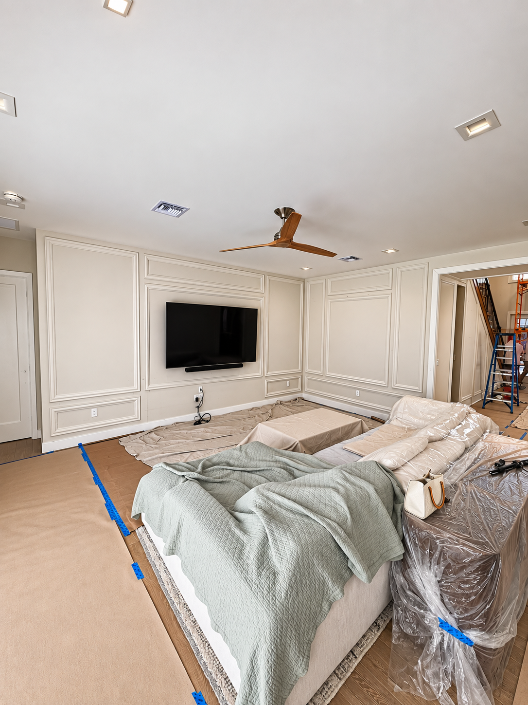 Bright bedroom with a large bed, wall-mounted TV, and ornate white paneling, covered in protective plastic and tape.
