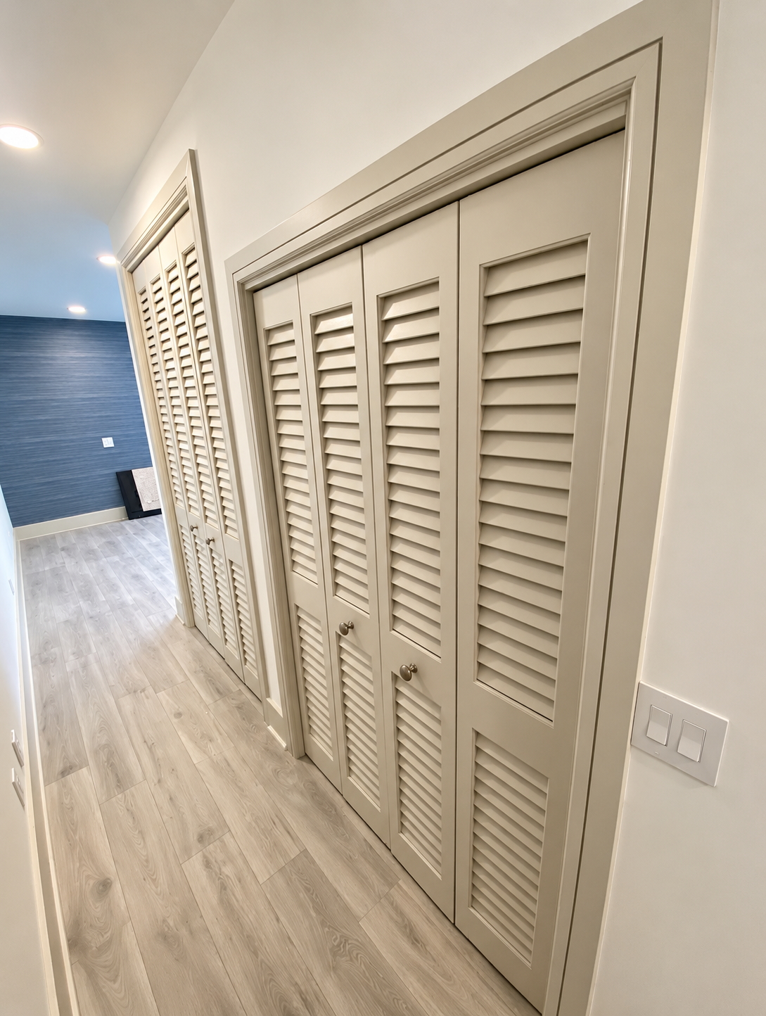 Narrow hallway with white louvered closet doors and light wood flooring, leading to a blue-tiled area
