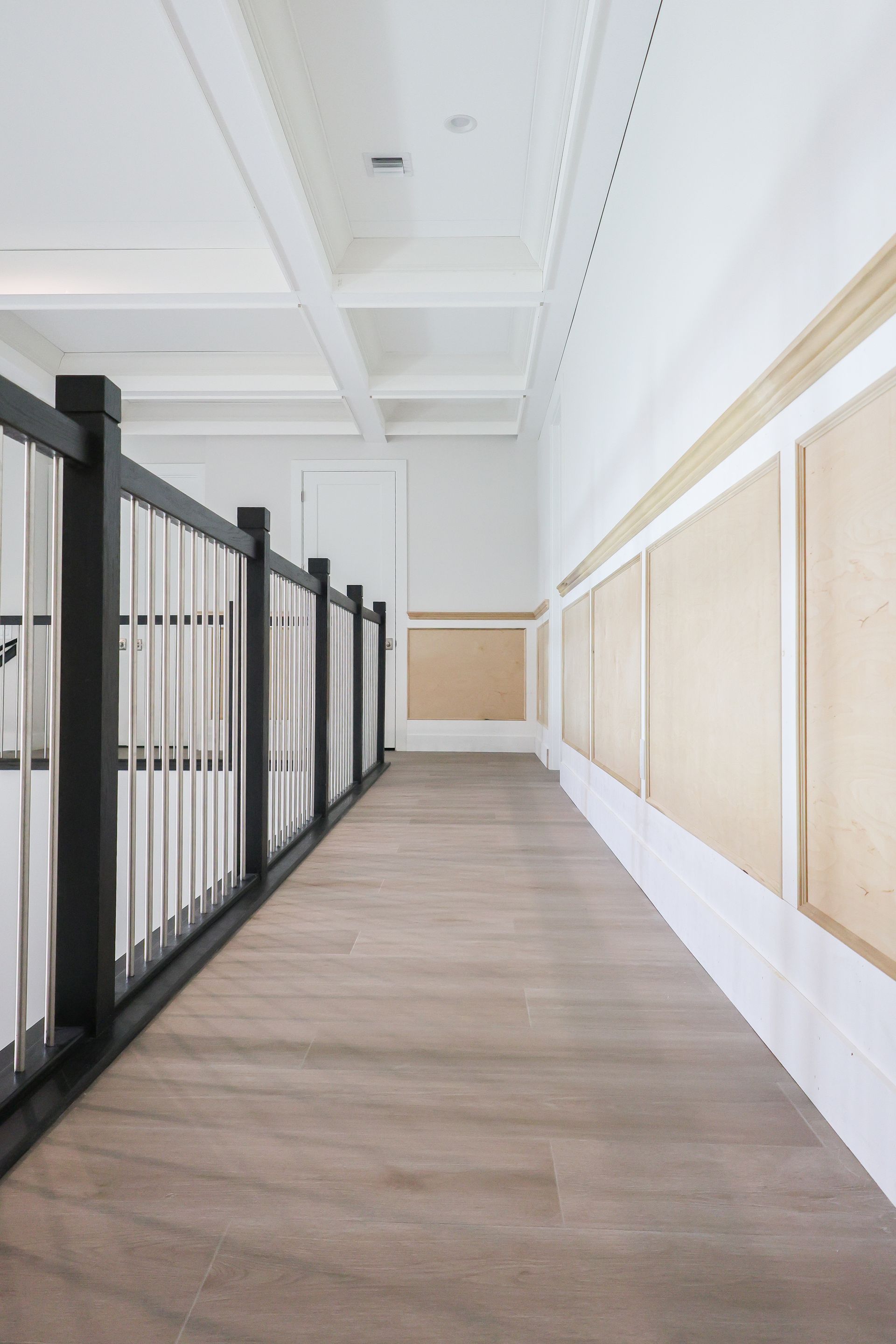 Empty hallway with black railings, light wood floor, and white paneled walls