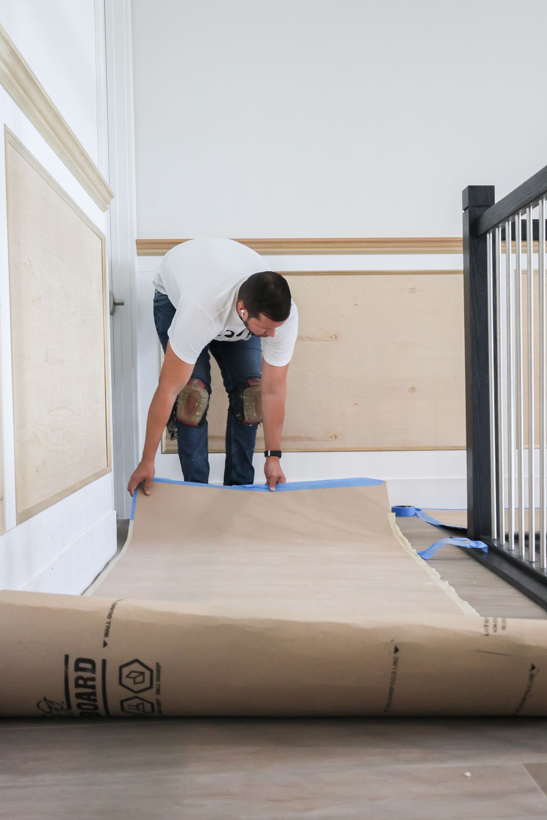 Man rolling out a large tan carpet in a hallway with white walls and a black railing