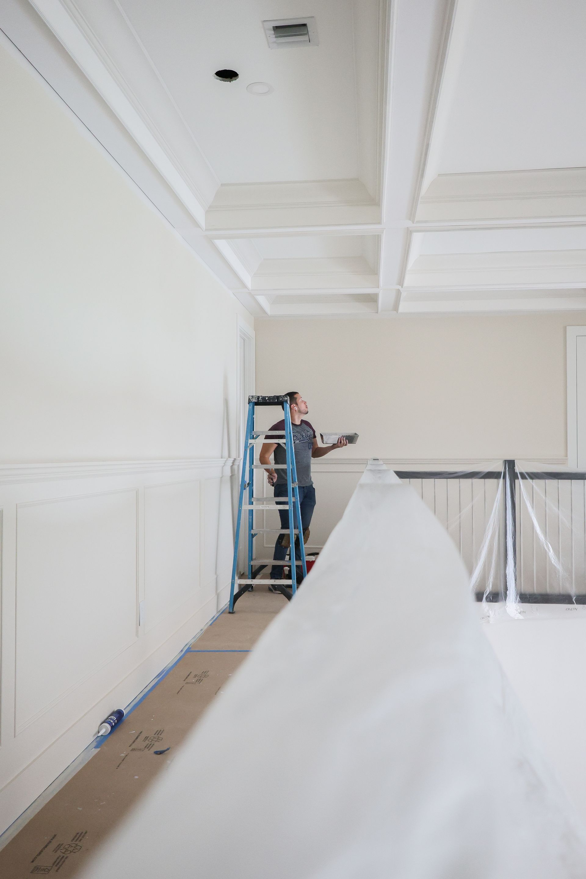 Person on ladder painting a white room with covered furniture and plastic sheeting