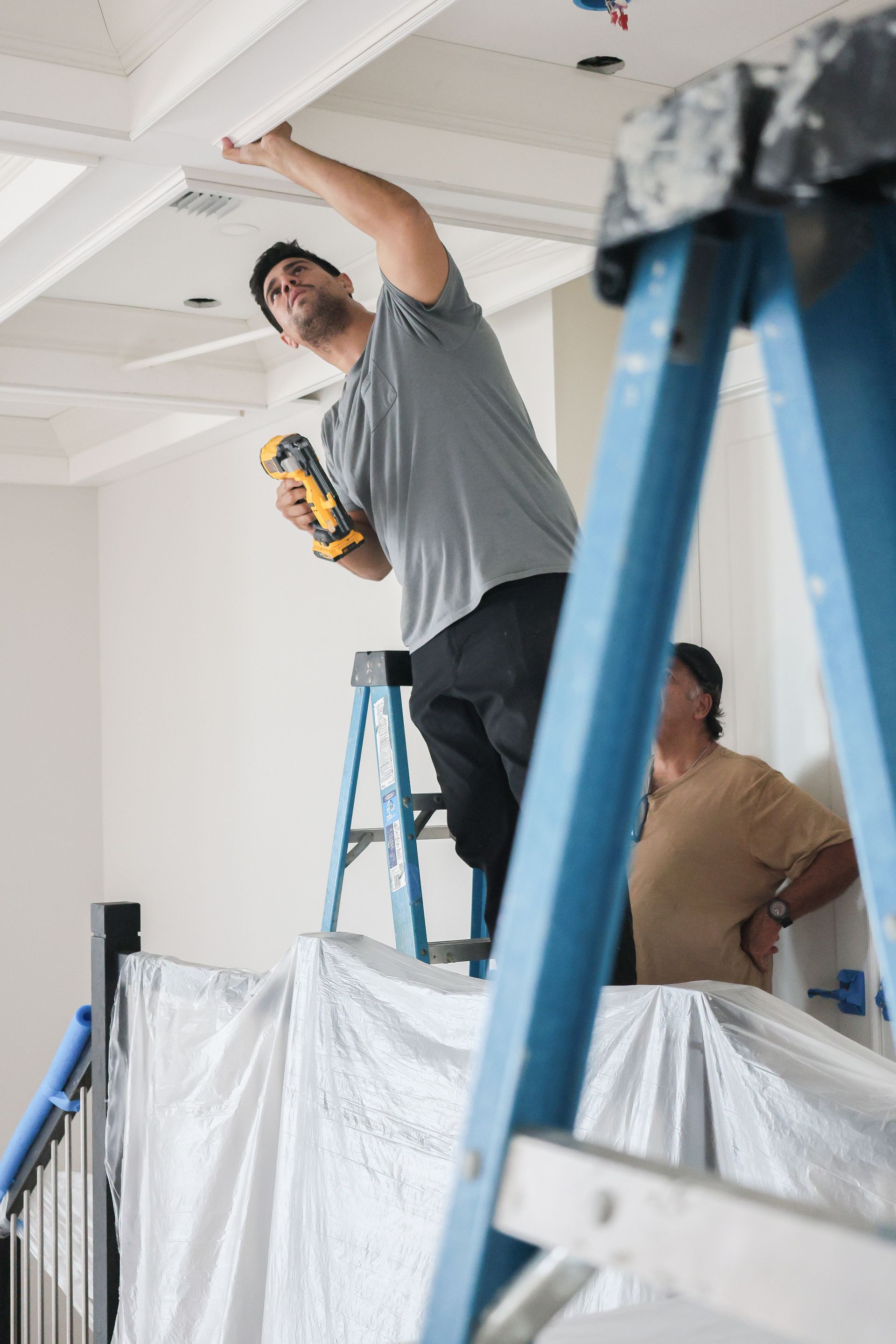 Workers painting a ceiling in a room, one standing on a ladder with a drill and another behind plastic sheeting.