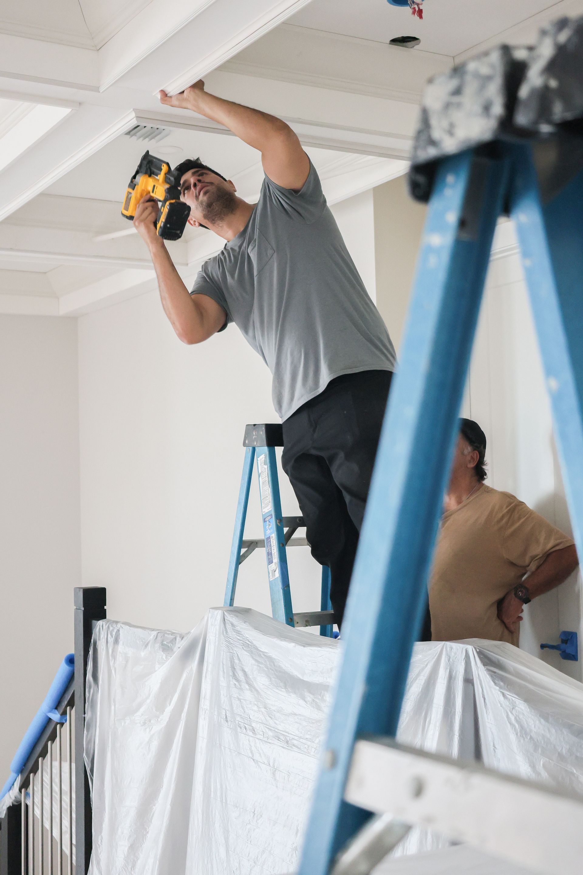 Worker on a step ladder installing ceiling trim while another person stands nearby under plastic-covered furniture.