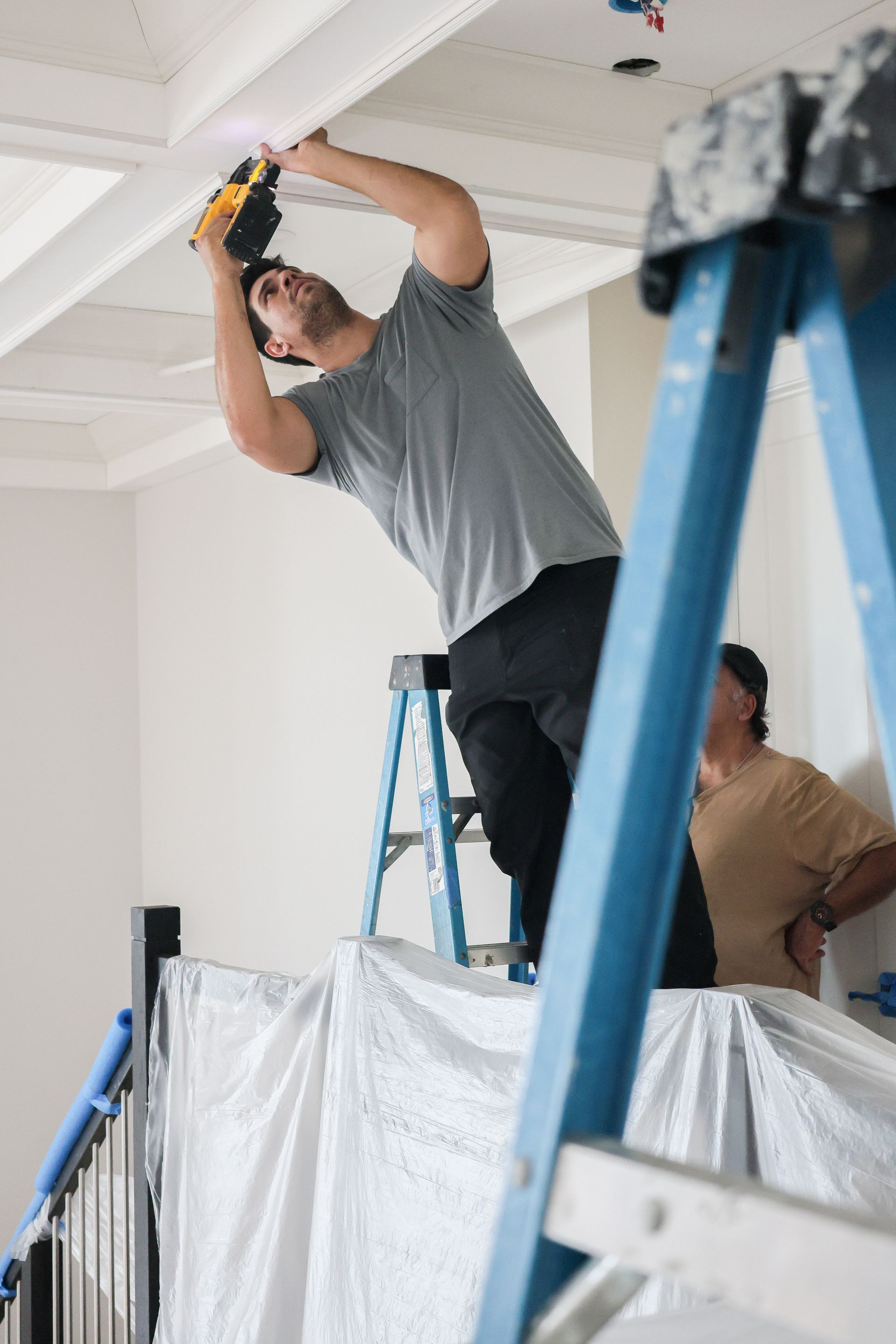 Two workers installing a ceiling light on a ladder, with protective plastic sheeting below.
