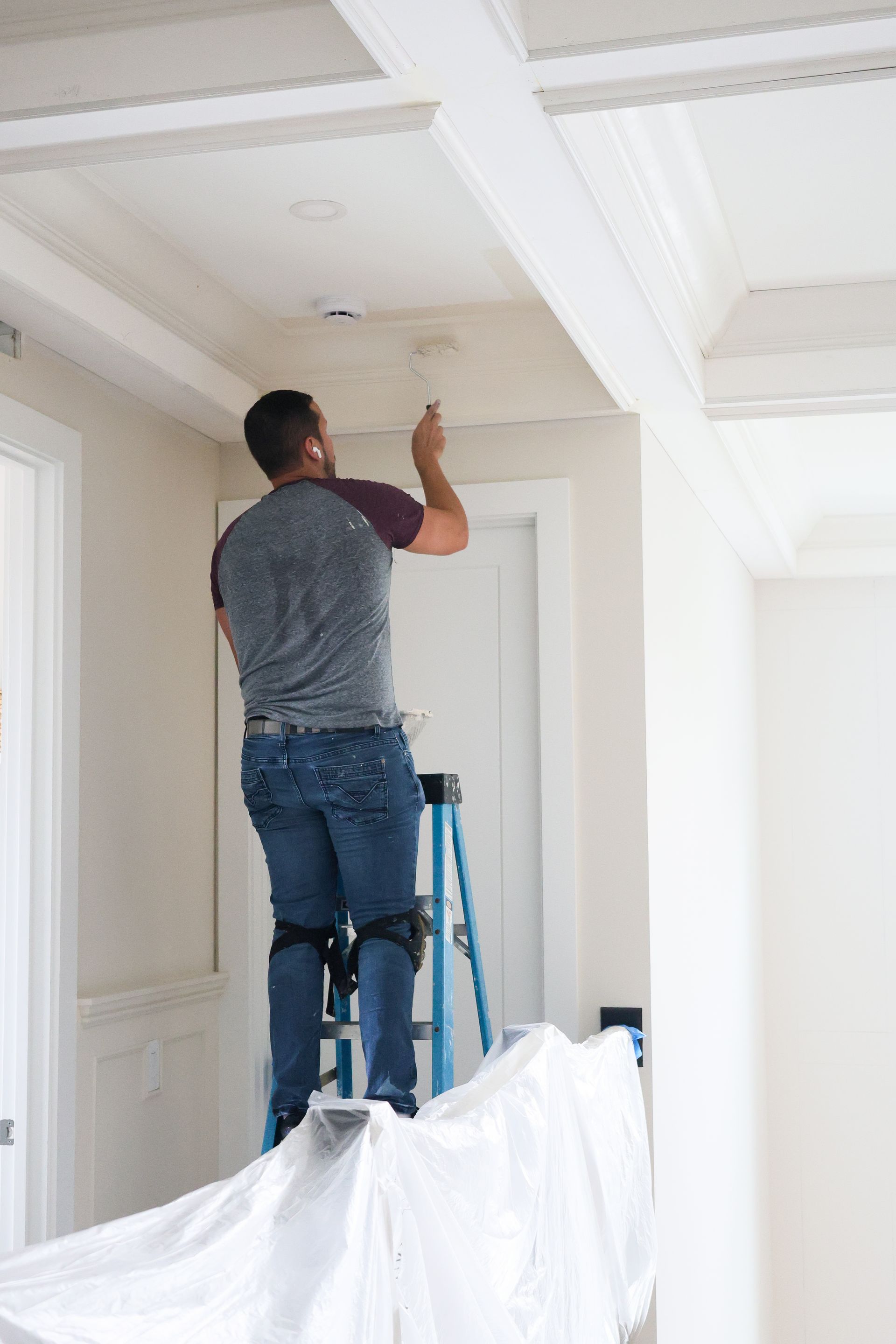 Person painting a white ceiling while standing on a ladder indoors.