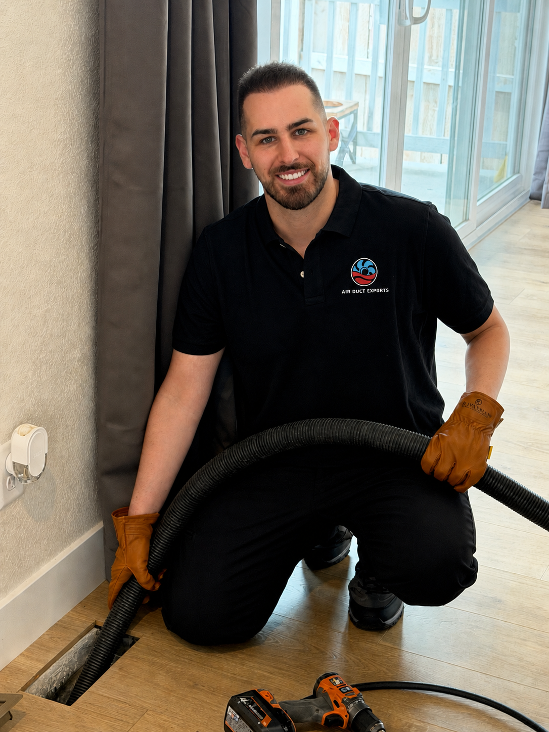 Worker kneeling with a vacuum hose, cleaning vent beside a window in a hallway.