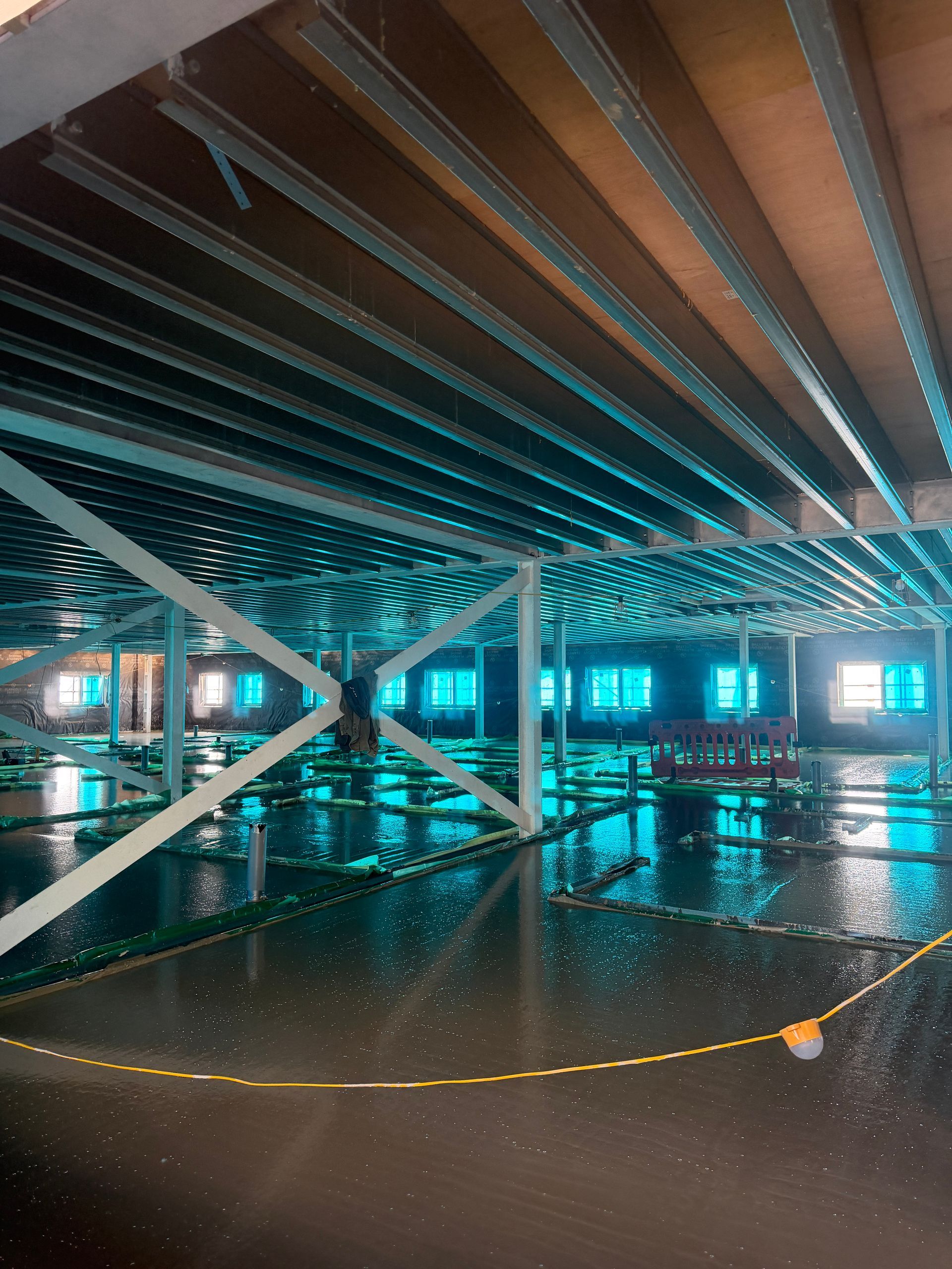 Flooded undercroft with steel beams, wet floor, and turquoise-lit windows reflected in standing water