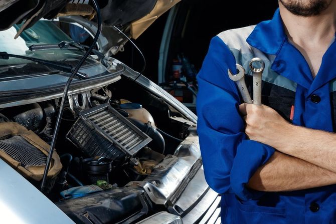 Meccanico in tuta blu, con in mano delle chiavi inglesi, in piedi vicino al motore aperto di un'auto.