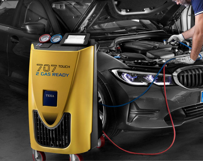 Meccanico controlla l'aria condizionata dell'auto con indicatori, vano motore visibile. Indicatori blu/rossi, guanti neri, parte anteriore dell'auto.