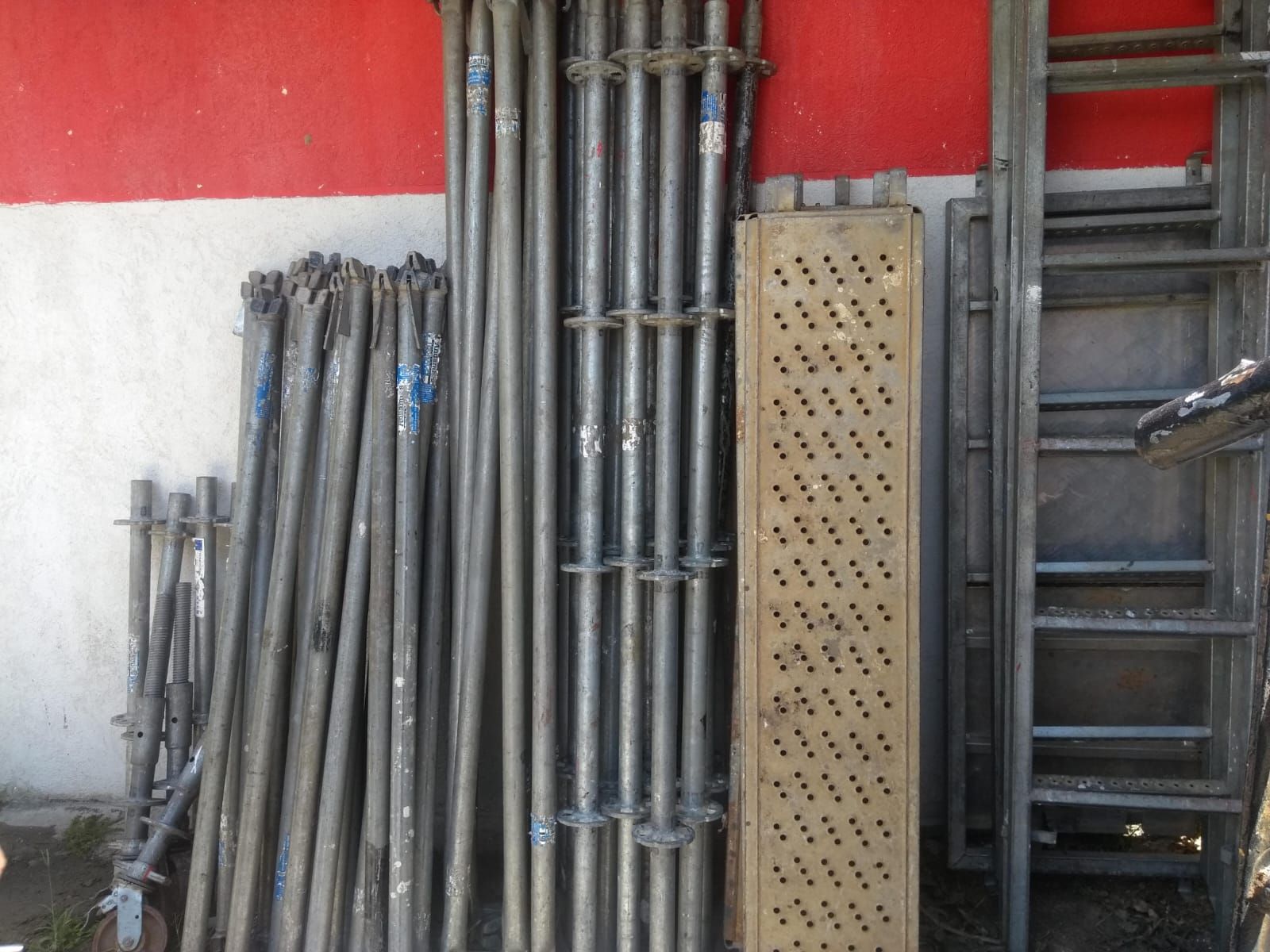 Andamios metálicos y escaleras apilados contra una pared roja y blanca en un área de almacenamiento.