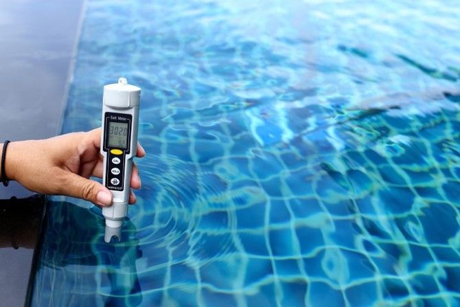 Hand holding a digital water tester over a blue swimming pool