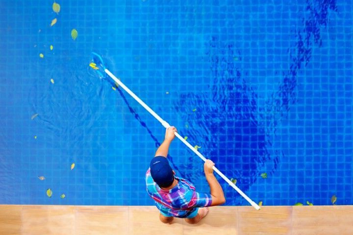 Person in blue cap cleans a blue-tiled wall with a long brush beside a pool