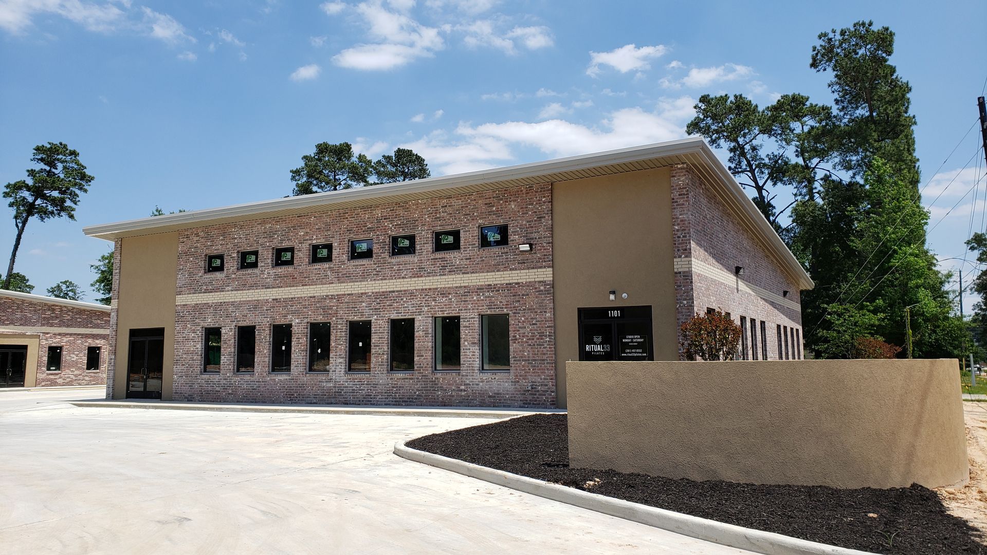 Modern brick building with small windows, a tan wall, and trees under a bright blue sky