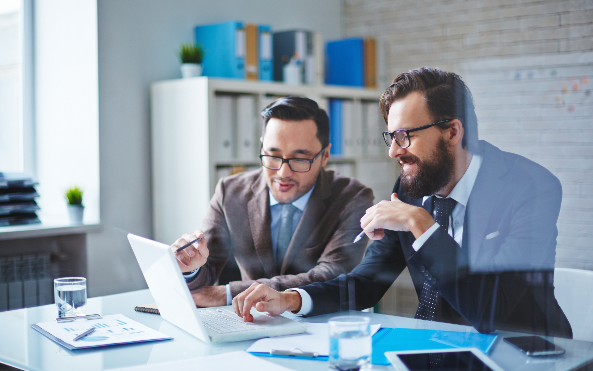 Two professionals discuss documents at a desk with a laptop in a bright office