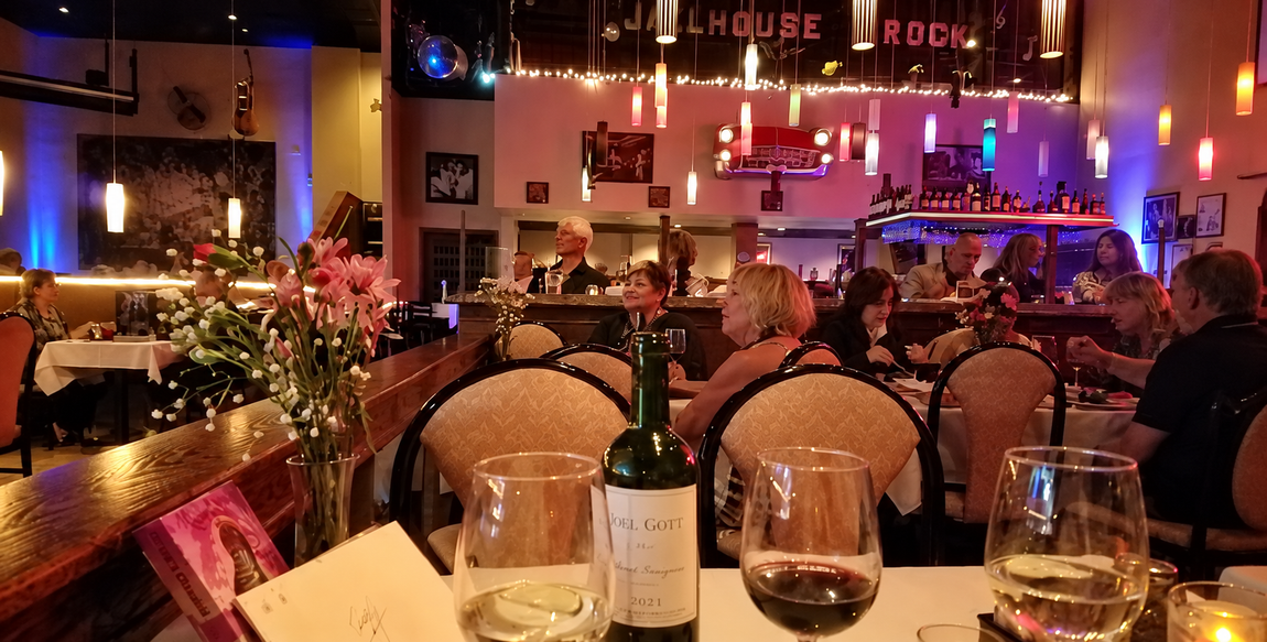 Restaurant table with wine glasses and bottle overlooking a warmly lit dining room and bar at night